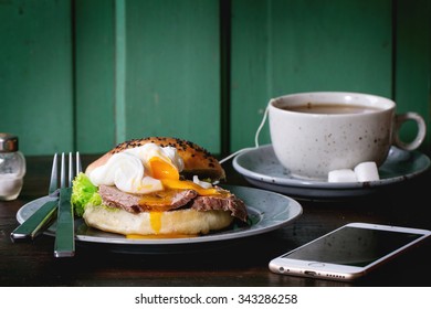 Cafe Breakfast With Homemade Sandwich With Baked Meat And Soft-boiled Egg, Cup Of Hot Pocket Tea And White Smartphone Over Dark Wooden Table With Turquoise Wall At Background. Dark Rustic Style.