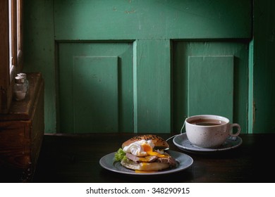 Cafe Breakfast With Homemade Sandwich With Baked Meat And Soft-boiled Egg And Cup Of Hot Pocket Tea Over Dark Wooden Table Near The Window With Turquoise Wall At Background. Dark Rustic Style.