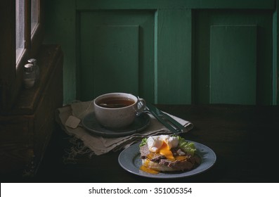 Cafe Breakfast With Homemade Sandwich With Baked Meat And Soft-boiled Egg And Cup Of Hot Pocket Tea Over Dark Wooden Table Near The Window With Turquoise Wall At Background. Dark Rustic Style.