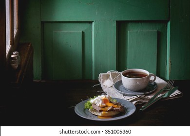 Cafe Breakfast With Homemade Sandwich With Baked Meat And Soft-boiled Egg And Cup Of Hot Pocket Tea Over Dark Wooden Table Near The Window With Turquoise Wall At Background. Dark Rustic Style.