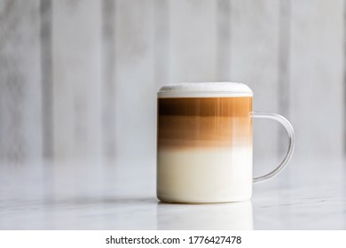 Cafe Latte Macchiato Layered Coffee In A See Through Glass Coffee Cup. The Cup Has A White Wooden Background.