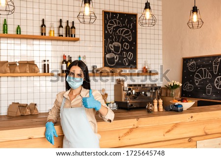 Cafe owner in medical mask and latex gloves looking at camera and showing like sign near table