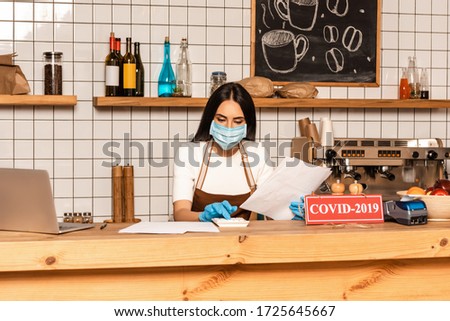 Cafe owner in medical mask with papers using calculator near laptop and card with covid-2019 lettering on table