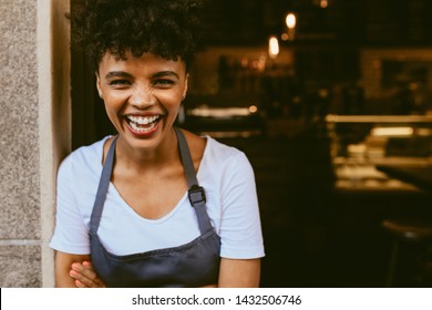 Cafe owner standing in the doorway of her coffee shop. Cheerful young female barista standing with her arms crossed at the door of a restaurant and looking at camera.