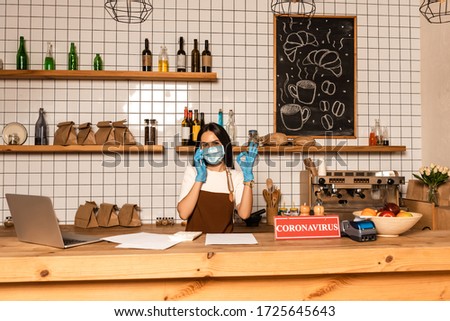 Cafe owner talking on smartphone and showing okay sign near table with laptop, papers, card with coronavirus lettering, payment terminal and bowl with fruits