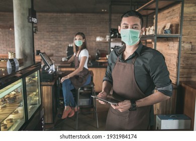 Cafe Owner Wear Face Mask At His Coffee Shop Using Tablet Pc. His Partner Sitting In A Background. Open For Business In New Normal Protocol