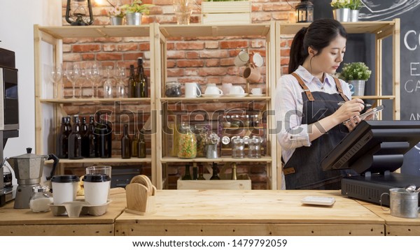 カフェ店員の女性が 顧客の注文を書いて請求カウンターに立っている ビンテージ喫茶店で 紙に書いたメモをペンで書いているレストランのオーナー エプロンのウェイトレスは仕事を集中して の写真素材 今すぐ編集