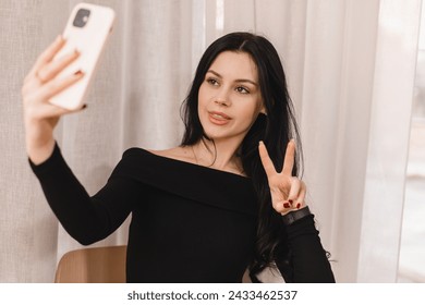 Cafe visitor demonstrating a V-sign to a frontal camera of a mobile phone while taking a picture of herself. Happy teen girl make selfie with peace gesture.