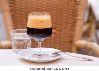 Caffe shakerato in a tall wine glass, in an outdoor italian cafe.  Cold shaked coffee with a foam made in a shaker with ice. Rattan chair on background. 