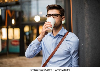 Caffee Break. Handsome Smart Casual Man With Eyeglasses And Leather Bag Having A Take Away Coffee In Urban Area Outside