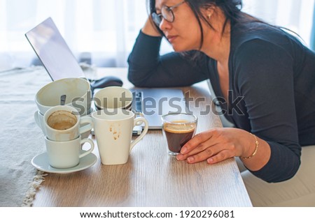 Caffeine addicted bad lifestyle concept. Young Asian woman holding a cup of coffee sitting tired with many empty cups of coffee and laptop on the desk. Focus on the hand and coffee.