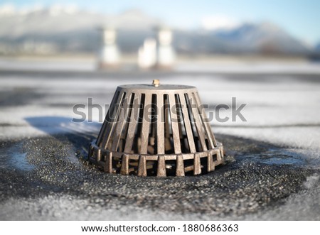 Caged roof drain with defocused vents and winter mountain background. Flat roof with exposed modified bitumen membrane. Inspect and clean roof drain system to prevent water damage. Selective focus.