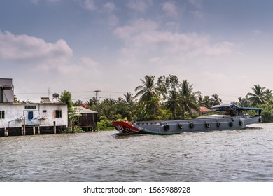 Cai Be, Mekong Delta, Vietnam - March 13, 2019: Along Kinh 28 Canal. Modern Gray Motorized Barge Sailing On Brown Water Under Blue Cloudscape. White House On Stilts And Green Foliage.