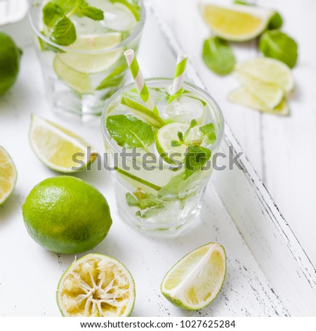 Caipirinha or mojito cocktail with lime, ice and mint leaves in beautiful glasses and cut green citrus on white wooden background. Summer alcohol drink.
