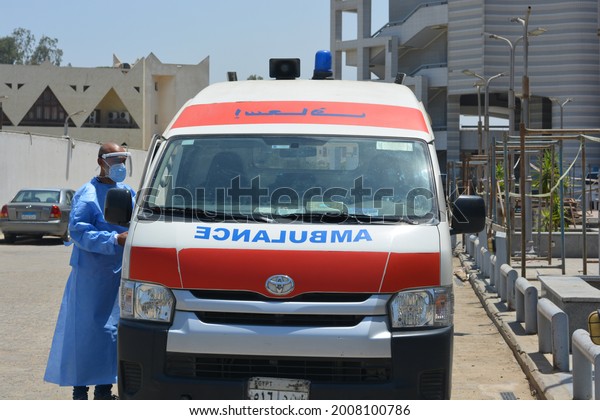 Cairo, Egypt, July 7 2021: Egyptian health worker\
in a blue protective gown, double face mask and face shield beside\
an equipped ambulance car ready to deal with covid-19 coronavirus\
emergency cases