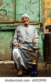 Cairo, Egypt - October 11, 2010: Friendly Old Egyptian Man Wearing A Long Gray Robe, A Jellabiya, Sitting, Laughing At Graffiti Wall Outdoor Street Cafe Smoking Sheesha Water Pipe In Islamic Cairo