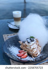 Cake Napoleon sprinkled with powdered sugar, decorated with berries and cotton candy.  In the background, a cup of latte and the sea. - Shutterstock ID 2258705723
