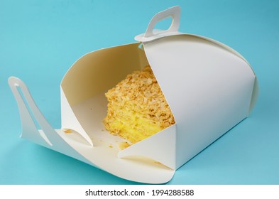 The Cake Is Packaged In A White Paper Box On A Light Blue Background. Open Kraft Packaging With A Piece Of Sweet Layer Cake. Eco-friendly Lunch Box. Food To Take Away Concept.