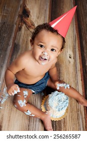 Cake Smash!  Adorable baby boy sitting on the floor, looking up, and enjoying a birthday cake.  