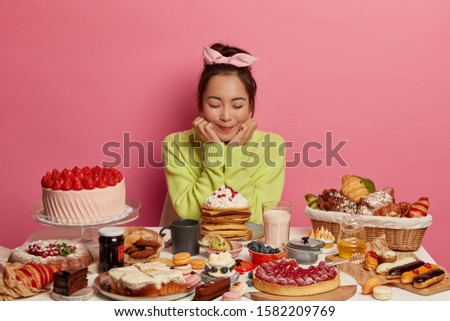 Cake tasting. Delighted Asian woman wears headband and green jumper, holds chin, has good appetite, eats sweet food, fruit cakes, comes on birthday party, isolated on pink wall, eager to eat