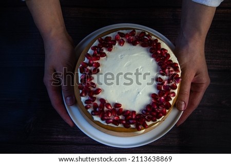 A cake of three layers, consisting of cakes and white cream, decorated with pomegranate on top, is held in the hands of a cook on a dark background, top view. High quality photo
