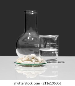 Calcium Carbonate Chip In Chemical Watch Glass Place Next To Crystal Clear Liquid In Beaker And Flat Bottom Flask (Borosilicate Glass). Closeup Chemical Ingredient On White Laboratory Table.