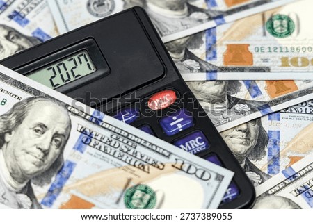 Calculator shows 2027 while surrounded by numerous hundred-dollar bills, emphasizing financial calculations and currency management in a detailed close-up