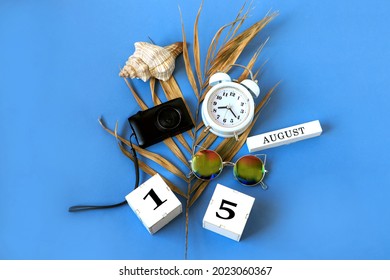 Calendar for August 15 : the name of the month of August in English, cubes with the number 15, a dry palm leaf, a shell, a camera, a watch, sunglasses on a blue background, a top view