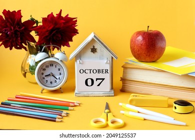 Calendar for October 7 : decorative house with the name of the month in English and the numbers 07, bouquet of dahlias, books, notebooks, pencils, alarm clock, red apple , yellow background