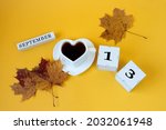 Calendar for September 13 : the name of the month in English, cubes with the number 13,a white heart-shaped cup on a saucer with tea, maple leaves, yellow background, top view