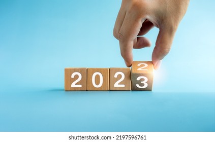 The Calendar Year 2022 Changed To 2023, Success Concept. Wooden Cube Blocks Turning By Hand For The Transition From 2022 To 2023, Preparation For Merry Christmas And Happy New Year On Blue Background.