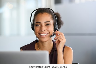 Call center agent with headset working on support hotline in modern office. Young african american agent in conversation with customer over headset looking at camera. Portrait of black girl working. - Shutterstock ID 2234751765