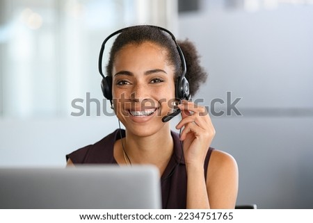 Call center agent with headset working on support hotline in modern office. Young african american agent in conversation with customer over headset looking at camera. Portrait of black girl working.