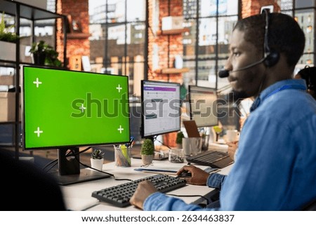 Call center worker in office solving customers issues using green screen PC. African american man in customer support job in front of chroma key computer display using headset to talk with clients