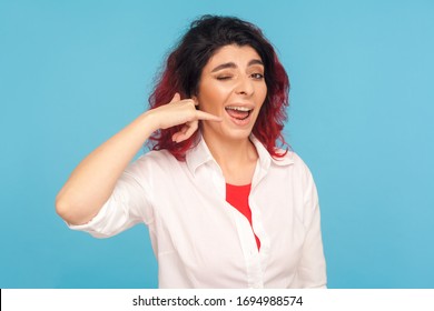 Call me back! Portrait of happy woman with fancy red hair making telephone gesture near head and winking playfully, waiting for call, wants to talk on phone. studio shot isolated on blue background