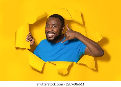 Call Me. Happy Black Man Making Contact Sign Gesture With Hand, Posing In Ripped Torn Yellow Paper Background. Communication And Mobile Cellular Connection Concept