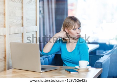 Call me! Portrait of cute young girl with blonde hear in blue t-shirt are sitting in the cafe and flirting with you, showing gesture call sing with fingers and making kiss with lips,looking at camera