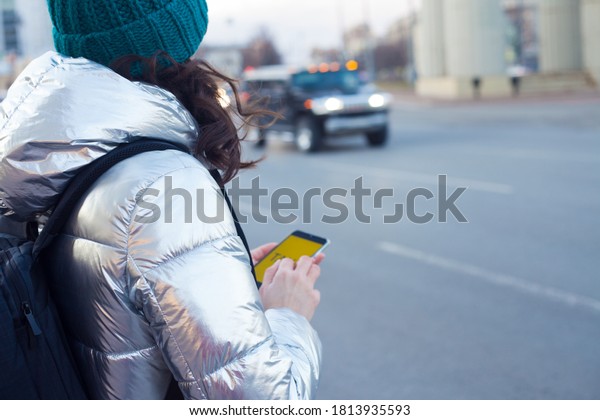 Call a taxi using a mobile app, a young woman in a\
down jacket and hat is standing by the road and waiting for a car.\
Autumn or winter city