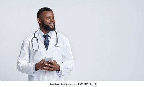 Call Your Doctor During Quarantine. Happy African Doctor With Mobile Phone Looking At Empty Space Over White Studio Background, Panorama