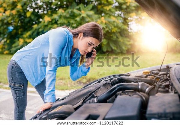 Calling Emergency\
Service. Car problems. Young woman using mobile phone while looking\
at broken down car on street. Woman caling autoservice because of\
car problem