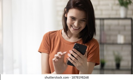 Calling to someone especial. Happy millennial woman stand at living room hold phone in hands dial number on touchscreen. Cheerful young lady surfing internet choosing name from contact list on cell