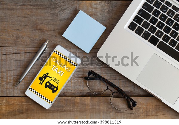 Calling Taxi message on a mobile phone screen. Top\
view of workplace with open laptop, glasses, pen and post-it over\
wooden table.