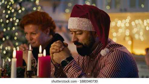 Calm Adult Bearded Man In Shirt And Santa Claus Hat Giving Prayer With Family While Having Festive Dinner On Xmas