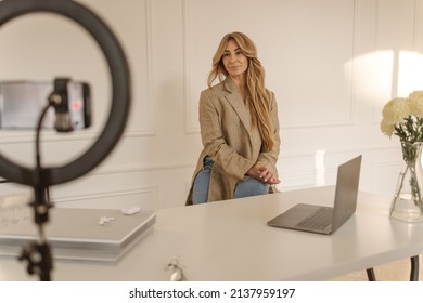 Calm Adult Caucasian Woman Looking At Phone Camera With Ring Light While Sitting In Office. Long-haired Blonde Wears Jacket And Jeans. Modern Gadgets Concept