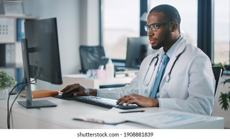 Calm African American Family Medical Doctor In Glasses Is Working On A Desktop Computer In A Health Clinic. Physician In White Lab Coat Is Browsing Medical History Behind A Desk In Hospital Office.