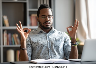 Calm African American man sit at desk meditating relieving negative emotions breathing fresh air, relaxed biracial male distracted from work practice yoga with mudra hands, stress free concept
