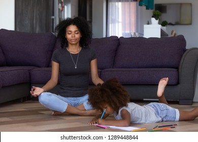 Calm african american mom doing yoga while quiet little mixed race kid daughter drawing on warm floor at home, black mother meditating for stress relief relaxing in living room with child daughter