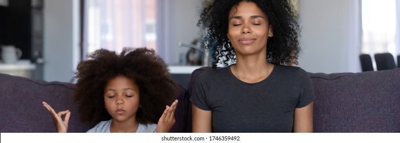 Calm African mother her funny little daughter sit on couch do meditation practice together at home. No stress, teach kid healthy life habits concept. Horizontal photo banner for website header design