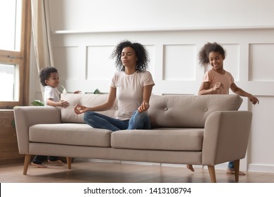 Calm African Single Mom Doing Yoga Exercises Sit On Sofa Reducing Stress With Active Noisy Kids Playing At Home, Mindful Black Parent Mother Meditating Relaxing With Children Running In Living Room