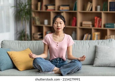 Calm asian lady sitting on comfy sofa and meditating with closed eyes at home, full length, copy space. Peaceful woman doing yoga in lotus pose, finding inner balance